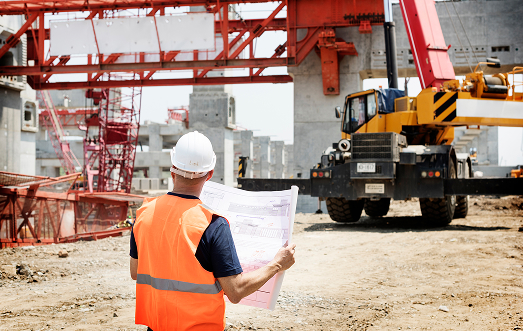 Site supervisor in a hard hat and safety vest reviewing construction plans at an industrial job site with a crane in the background